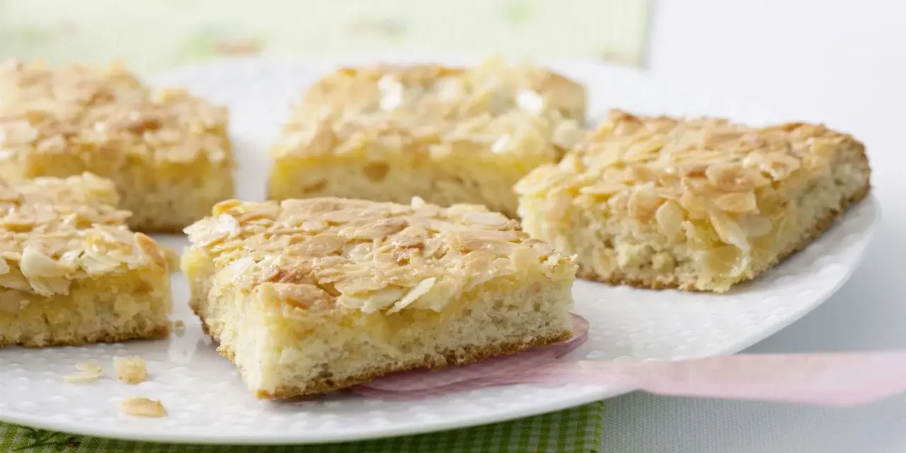 Saftiger mandelkuchen vom blech, goldbraun gebacken und mit knusprigen Mandelblättchen bestreut, liegt auf einer weißen Platte.