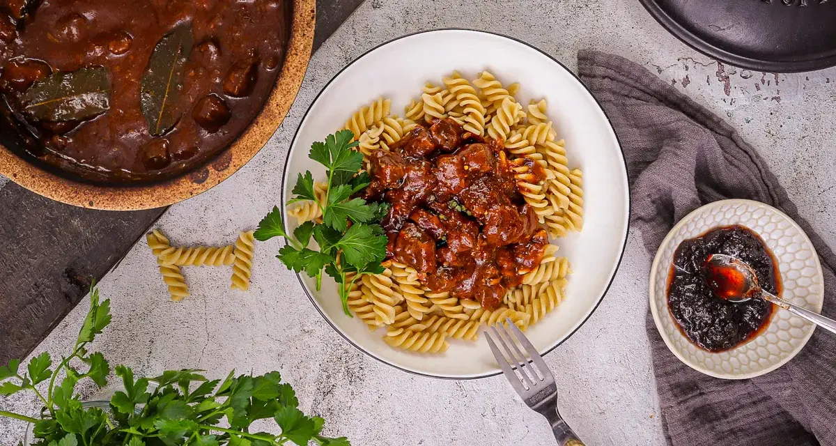 Herzhaftes Gulasch gemischt Rezept mit Fusilli, garniert mit Petersilie. Daneben ein kleiner Teller mit Preiselbeeren.