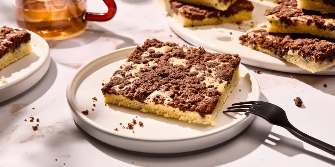 Ein Stück versunkener Streuselkuchen mit Schokostreuseln auf einem Teller, daneben eine Gabel und ein Glas Tee.