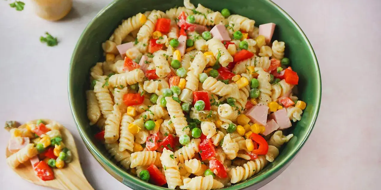 Ein bunter Nudelsalat mit Fleischwurst, Erbsen, Mais und Paprika in einer grünen Schüssel.