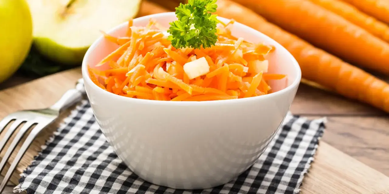 Frischer Karotten-Apfel-Salat in einer weißen Schüssel, garniert mit Petersilie. Im Hintergrund liegen ganze Karotten und ein halber Apfel.
