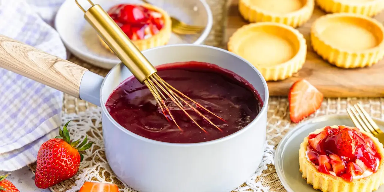 Frisch zubereiteter Tortenguss in einem Topf, bereit, um Obstkuchen zu verfeinern. Daneben liegen frische Erdbeeren und leere Tartelettes.