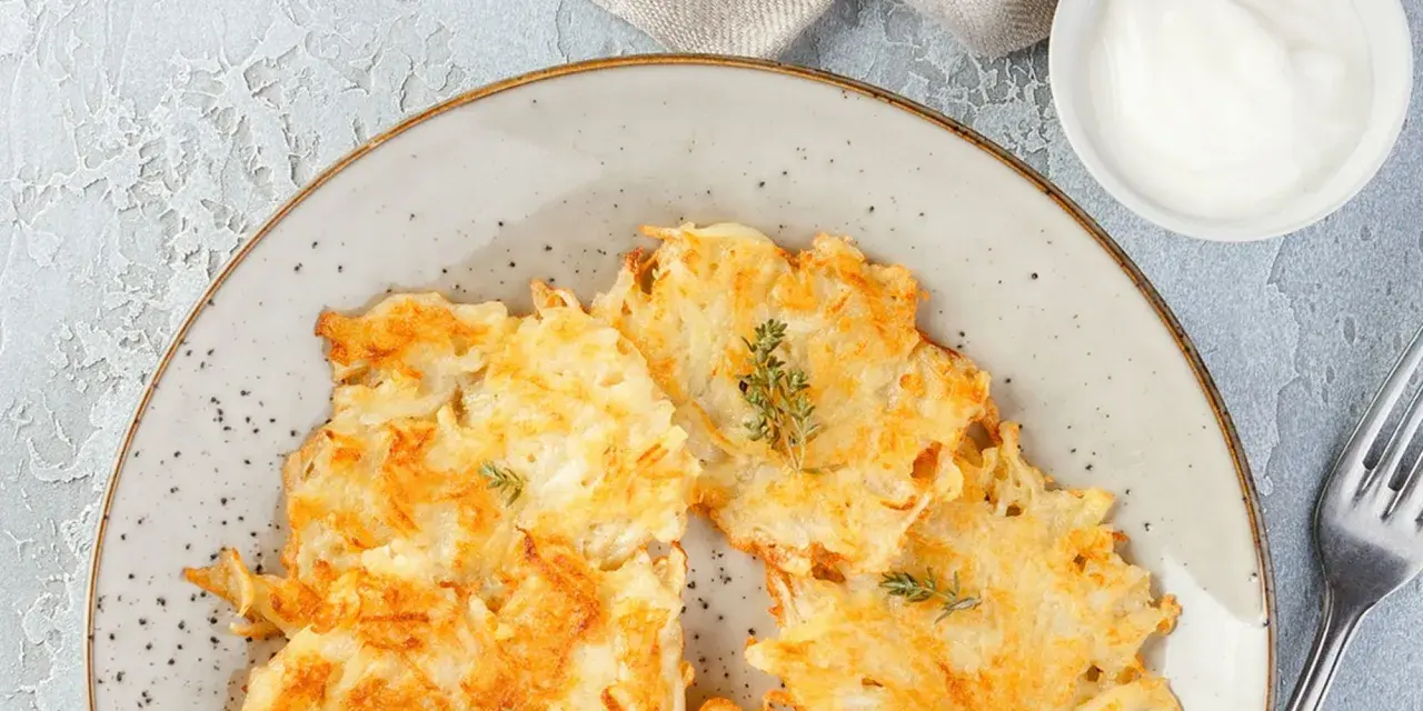 Knusprige Kartoffelpuffer, im Waffeleisen goldbraun gebacken, liegen auf einem Teller. Dazu ein Schälchen mit Sour Cream und ein Gabel.