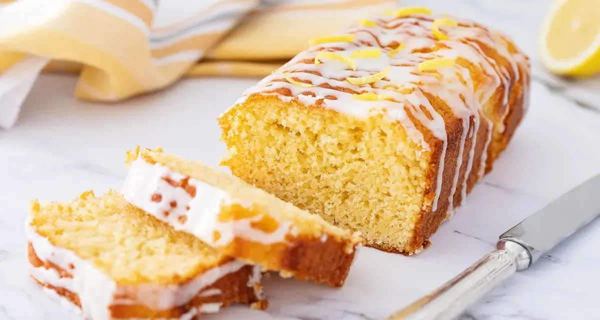 Saftiger zitronenkuchen mit Joghurt, überzogen mit Zuckerguss und garniert mit Zitronenschale. Ein Stück ist angeschnitten.