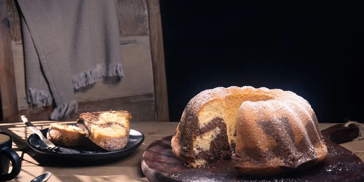 Ein fluffiger Marmorkuchen, bestäubt mit Puderzucker, liegt auf einem Holzbrett. Daneben zwei Stücke auf einem Teller.