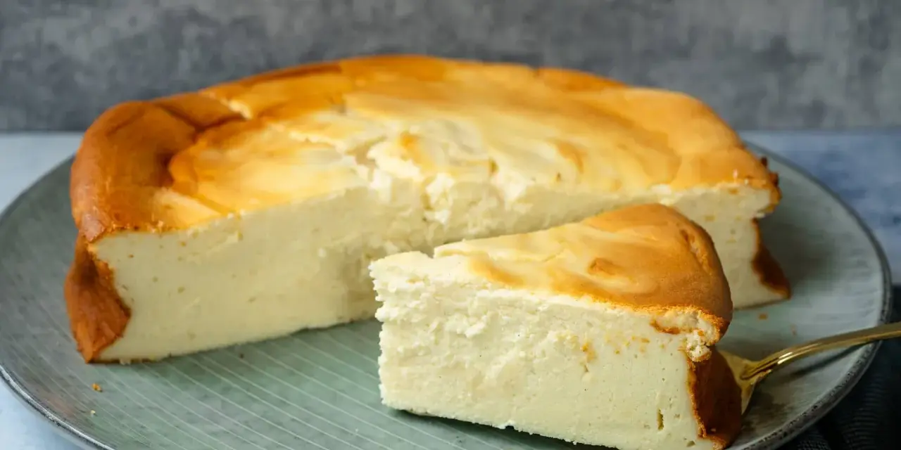 Ein saftiger Quarkkuchen ohne Boden, goldbraun gebacken, liegt auf einem Teller. Ein Stück ist bereits angeschnitten.