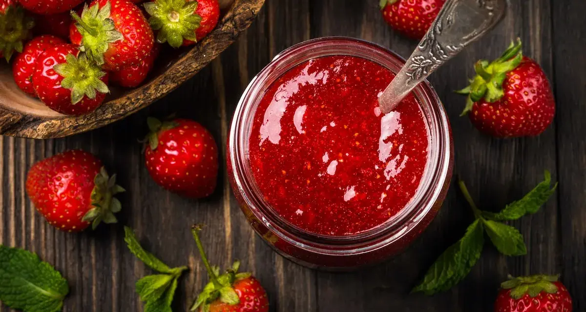 Frische Erdbeeren und ein Glas selbstgemachte Erdbeermarmelade, bereit zum Einkochen. Ein Löffel steckt in der Marmelade.