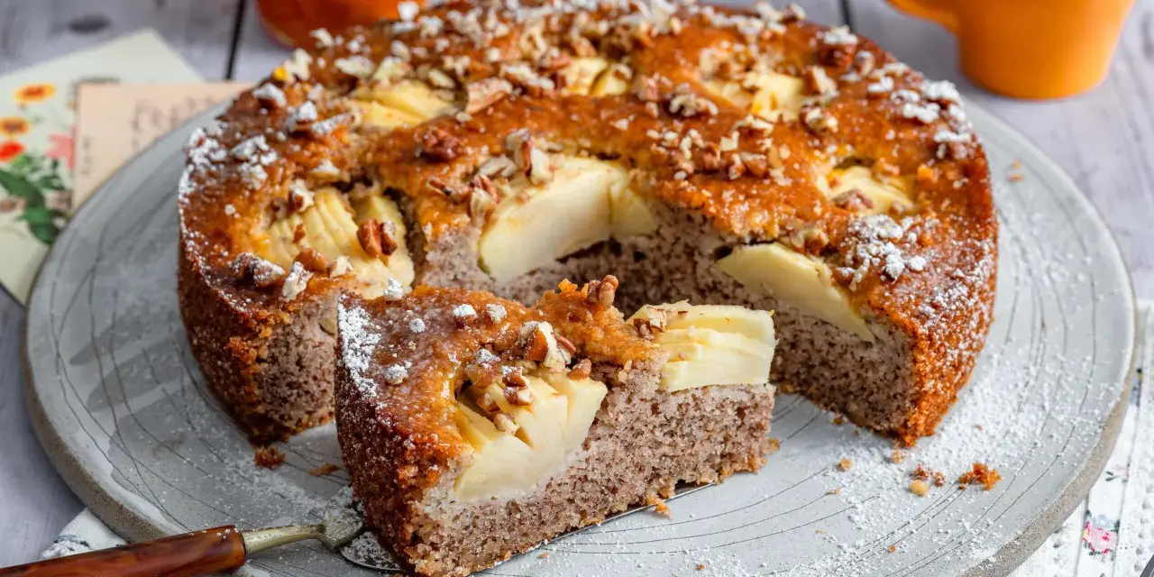 Ein saftiger Apfel-Birnen-Kuchen, bestreut mit Nüssen und Puderzucker, wird auf einem Teller serviert. Ein Stück ist bereits herausgeschnitten.