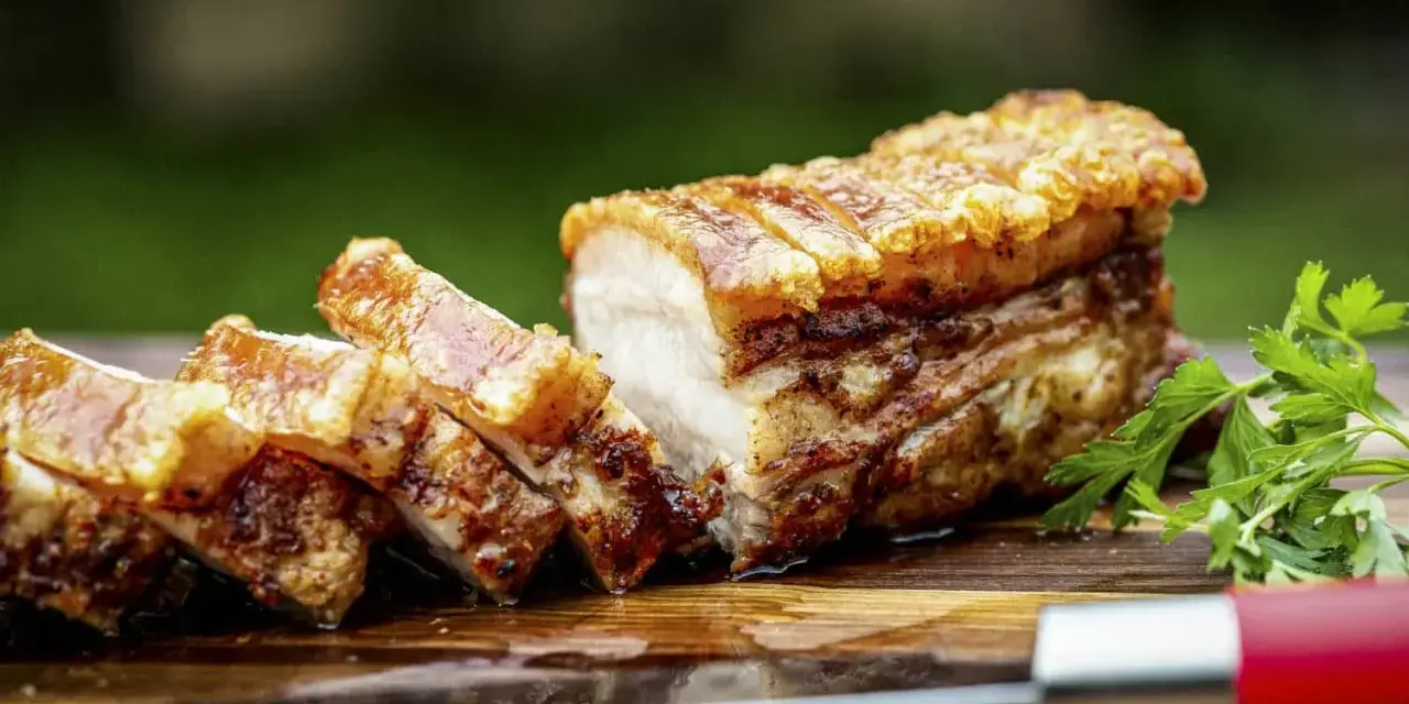 Knuspriger Schweinebauch, perfekt für ein köstliches schweinebauch rezept. Saftig, mit krosser Schwarte, auf einem Holzbrett mit Petersilie und einem Messer.