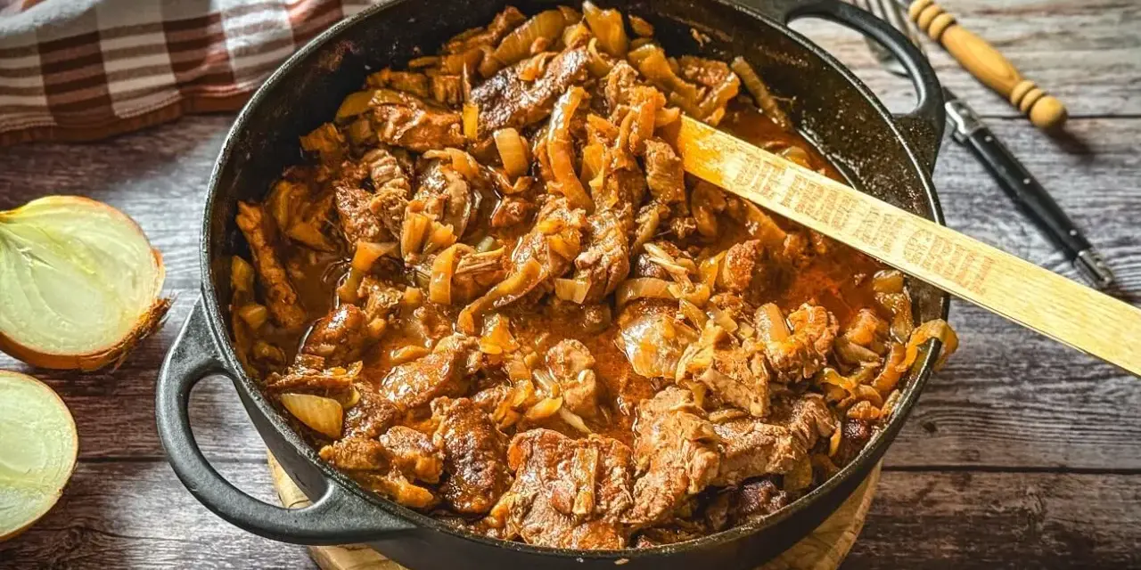 Herzhaftes Zwiebelfleisch-Rezept in Gusseisenpfanne, mit Zwiebeln und Fleischstücken in einer würzigen Sauce. Ein Holzlöffel mit der Aufschrift "Die Frau am Grill" liegt darin.