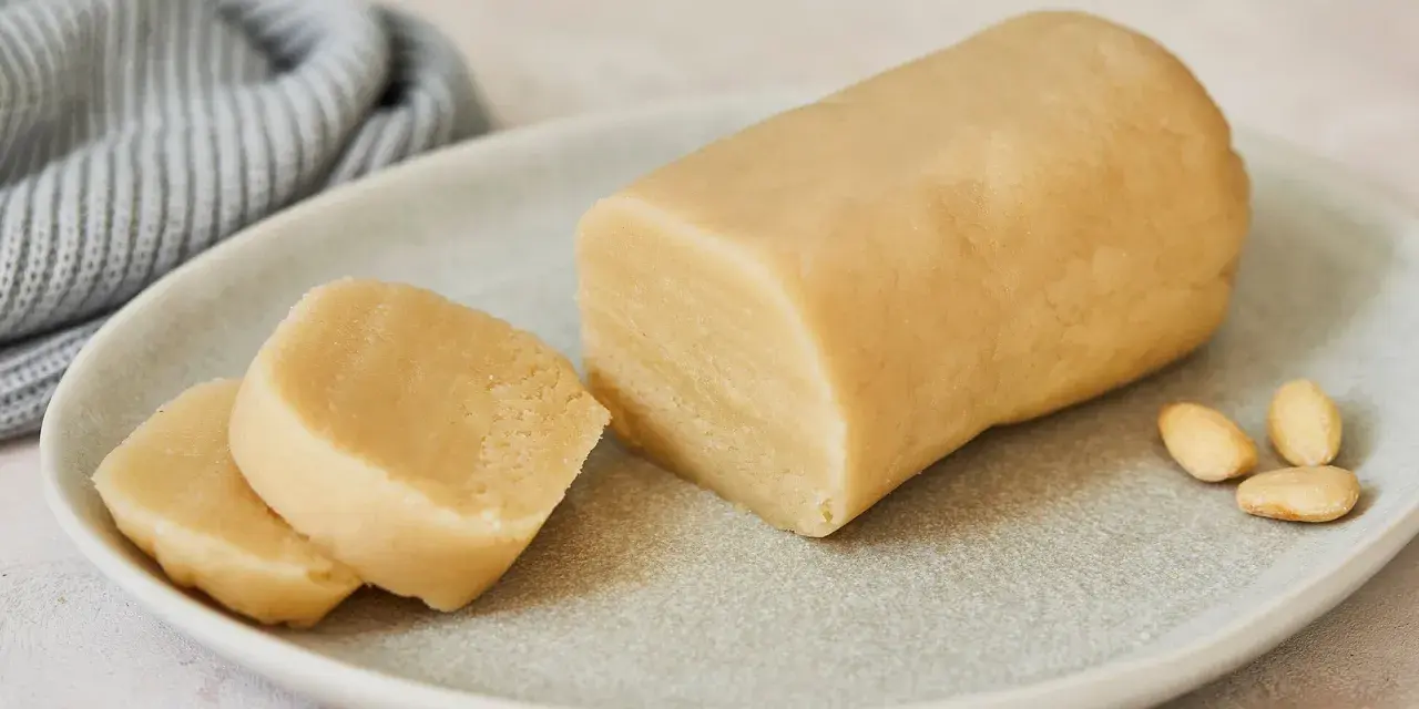 Frisch zubereitetes Marzipan zum **Marzipan selbst machen**. Ein Block und zwei Scheiben liegen auf einem Teller, daneben Mandeln.