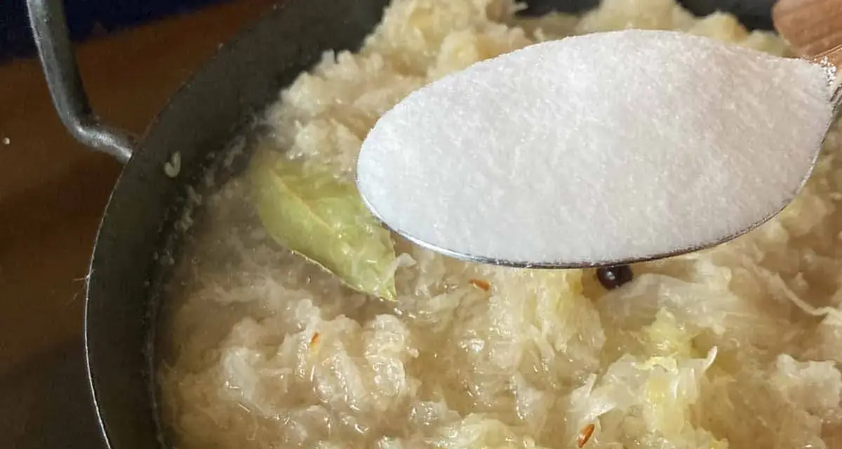 Zucker wird zu köchelndem Sauerkraut nach Omas Rezept gegeben. Ein Lorbeerblatt schwimmt im Topf.