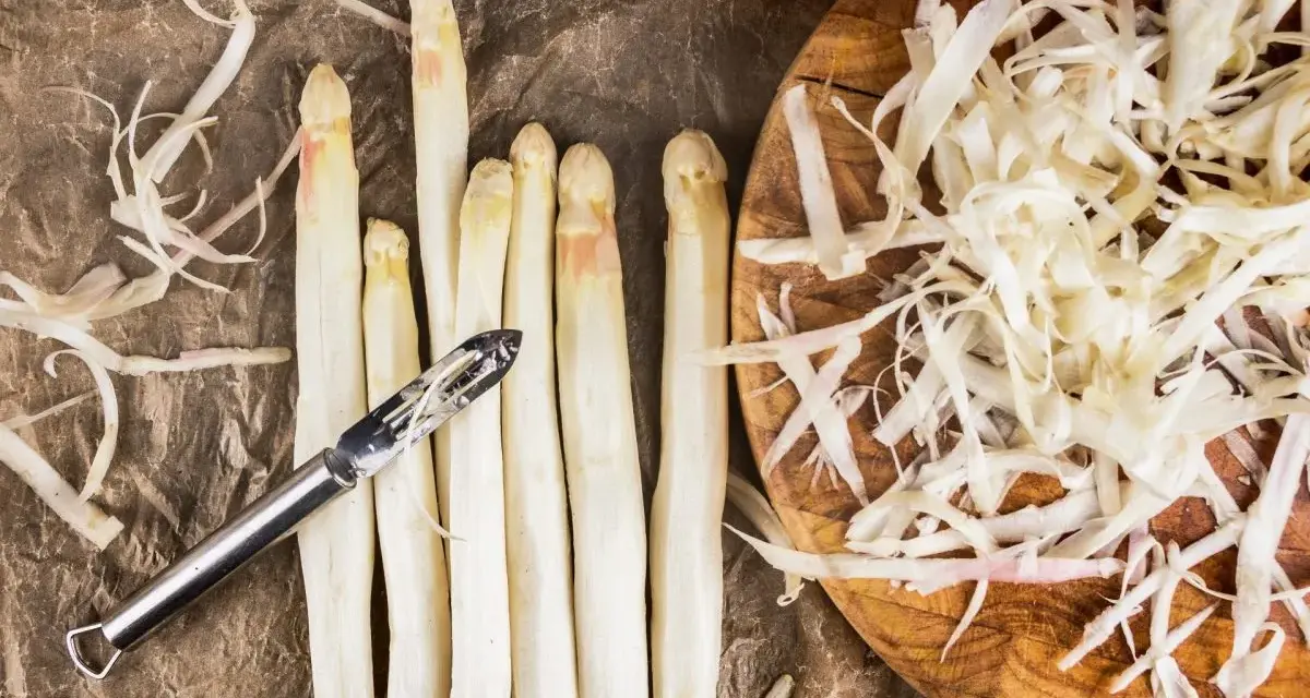 Frischer weißer Spargel wird geschält, die Spargelschalen liegen daneben. Ein Sparschäler liegt auf den Spargelstangen.