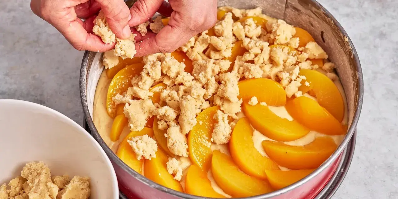 Hände verteilen Streusel auf einem Pfirsichkuchen in einer Springform.