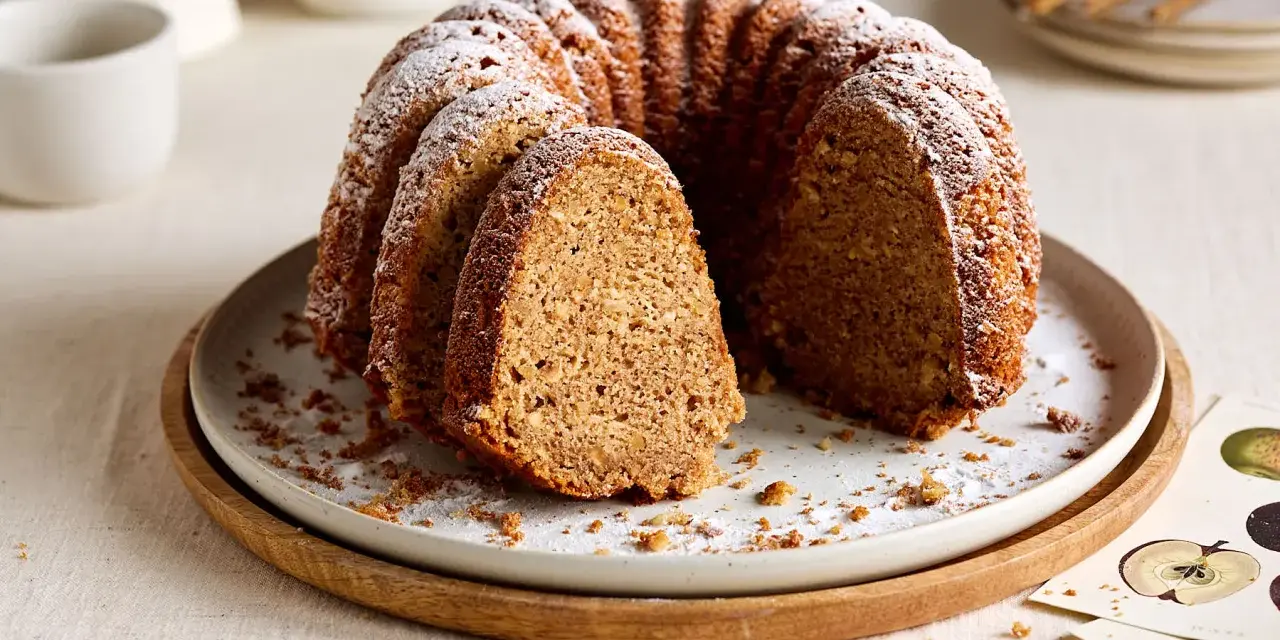 Ein saftiger Kuchen mit Dinkelmehl, bestäubt mit Puderzucker, liegt auf einem Teller. Ein Stück ist herausgeschnitten.
