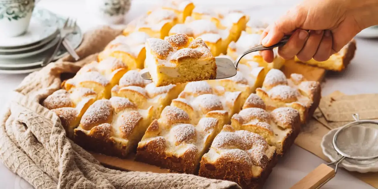 Ein Stück Spiegeleikuchen mit Quark wird aus einem Blechkuchen mit Puderzucker gehoben.