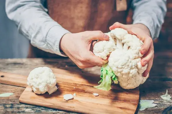 Hände zerteilen einen frischen Blumenkohl auf einem Holzbrett. So beginnt die Zubereitung eines gesunden Gemüses.