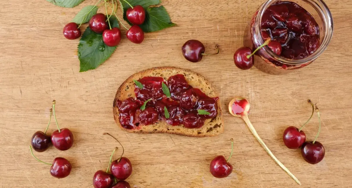 Frisches Brot mit Kirschmarmelade, garniert mit Minze. Ein Glas Kirschmarmelade und frische Kirschen daneben. Perfekt für ein Kirschmarmelade Rezept.