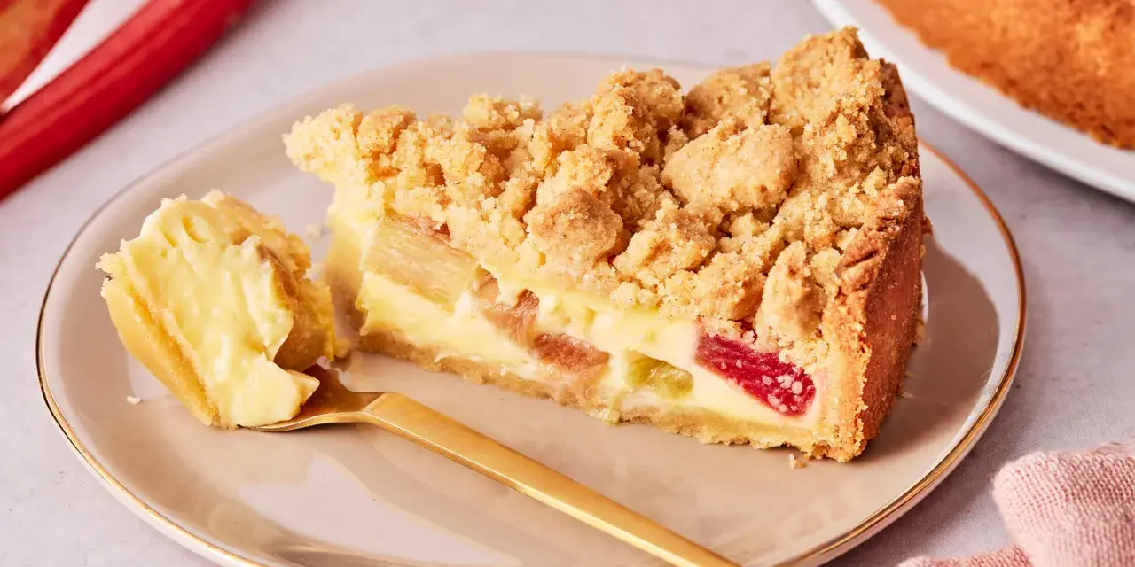 Ein Stück von Omas Rhabarberkuchen mit Pudding und Streuseln auf einem Teller, daneben ein goldener Löffel und rote Rhabarberstangen.