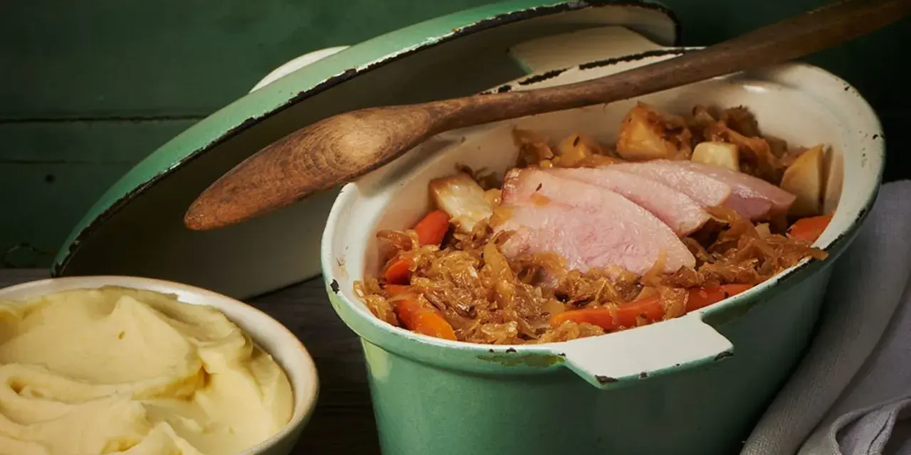Herzhafter Szegediner Gulasch mit zarten Fleischscheiben, Sauerkraut und Karotten in einem grünen Topf, daneben eine Schüssel cremiges Kartoffelpüree.