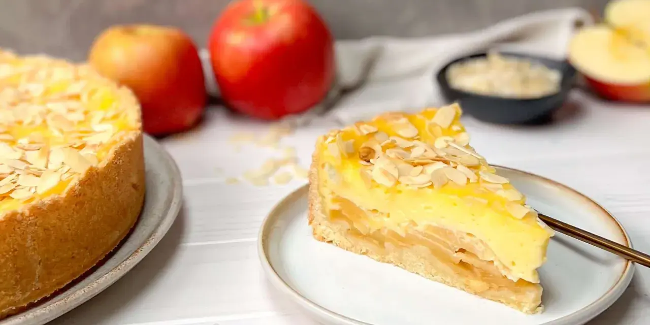 Ein saftiger Apfelkuchen mit Schmandguss, bestreut mit Mandelblättchen. Daneben liegen frische Äpfel.