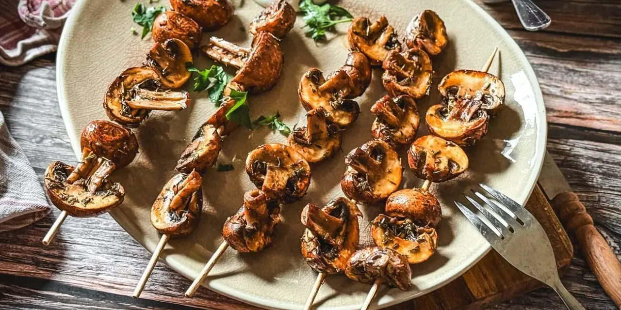 Gegrillte Champignons am Spieß, mit Kräutern verfeinert, auf einem Teller serviert. Ein Genuss für jeden Pilzliebhaber!