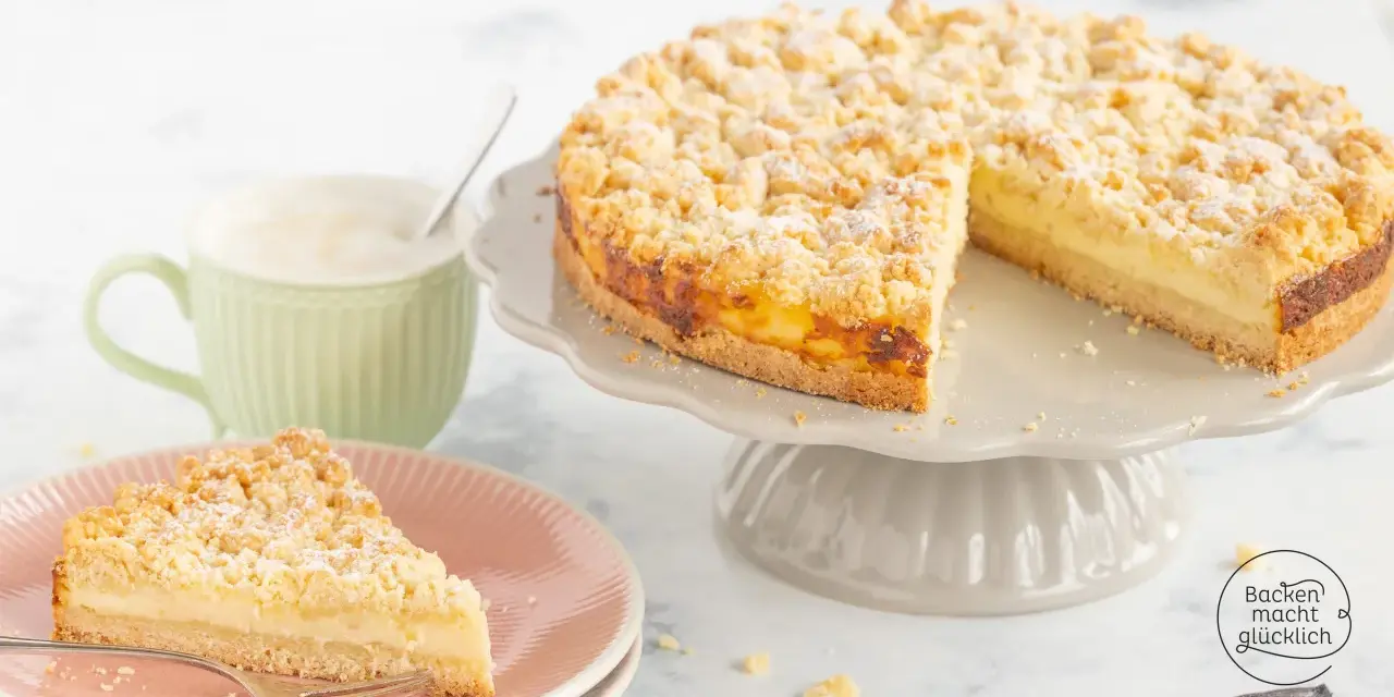 Ein saftiger Streuselkuchen mit Puddingfüllung, bestäubt mit Puderzucker. Daneben eine Tasse Cappuccino.