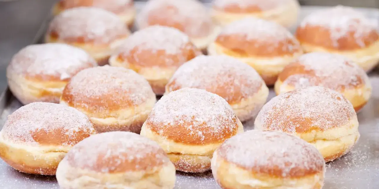 Eine Reihe von frisch gebackenen, mit Puderzucker bestäubten Berliner Pfannkuchen liegt auf einem Blech.