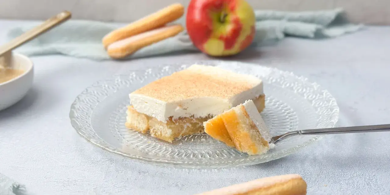 Ein Stück Apfel-Schmand-Kuchen aus der Springform, mit Löffelbiskuits und einem Apfel im Hintergrund.