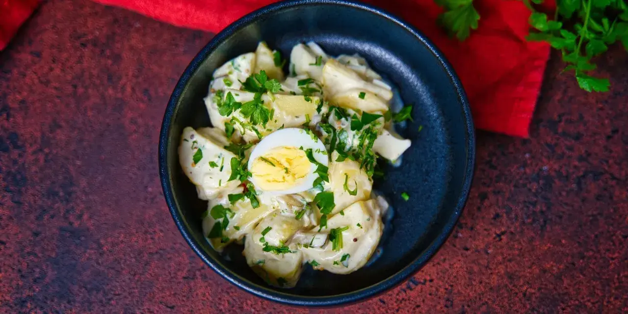 Ein cremiger Kartoffelsalat mit Mayo, garniert mit Petersilie und einem halben Ei, serviert in einer dunklen Schüssel.
