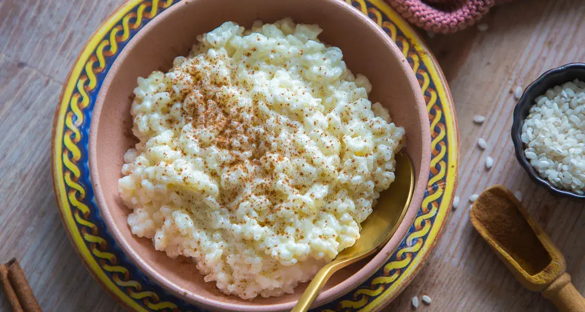 Cremiger Milchreis, bestreut mit Zimt, wartet in einer Schale auf sein Milchreis Rezept. Daneben liegen Reiskörner und Zimtstangen.