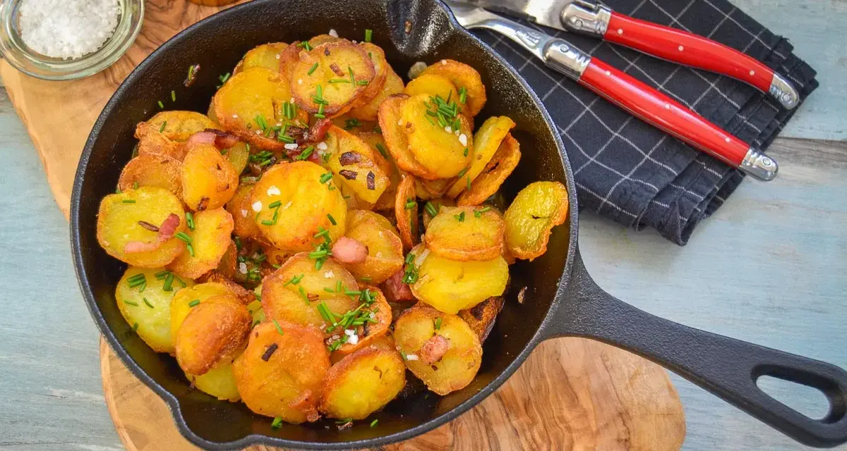 Bratkartoffeln mit Zwiebeln und Speck in einer Gusseisenpfanne, garniert mit Schnittlauch. Dazu grobes Salz und ein Messer-Gabel-Set.