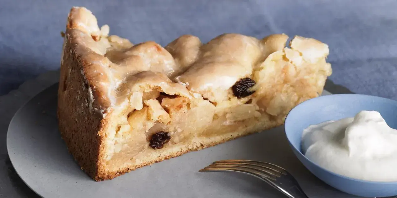 Ein Stück gedeckter Apfelkuchen mit Rosinen und Zuckerguss, serviert mit einem Klecks Sahne.