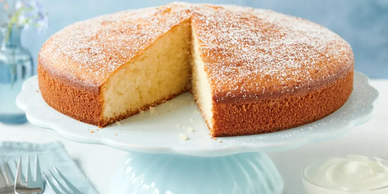 Ein **schneller saftiger Kuchen mit Joghurt**, bestäubt mit Puderzucker, auf einem hellblauen Tortenständer. Daneben eine Schale mit Sahne und Gabeln.