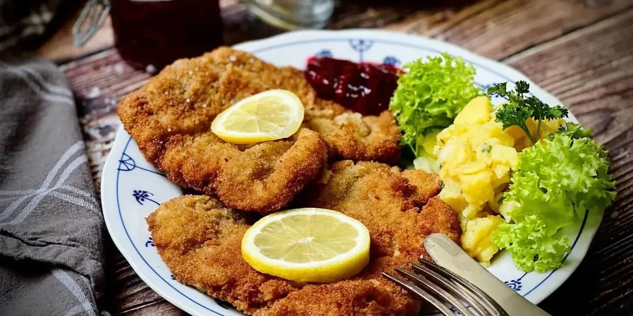 Knuspriges Wiener Schnitzel mit Zitrone, Preiselbeeren und Kartoffelsalat auf einem Teller.