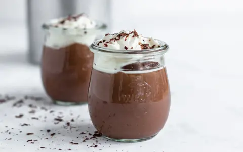 Zwei Gläser mit cremigem Schokopudding, gekrönt mit Schlagsahne und Schokostreuseln. Perfekt, um Schokopudding selber machen zu lernen!