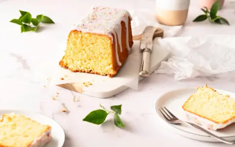 Saftiger zitronenkuchen kastenform mit Zuckerguss und bunten Streuseln, auf einem Marmorsockel, daneben ein Messer und ein Teller mit einem Stück Kuchen.