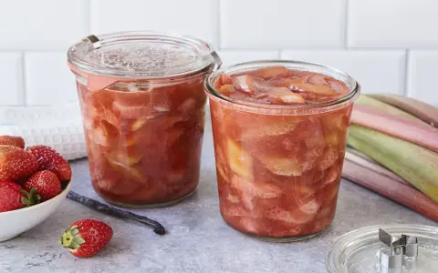Zwei Gläser mit leckerem Rhabarberkompott, daneben frische Erdbeeren und Rhabarberstangen.