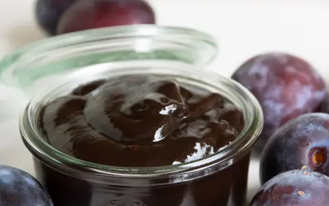 Ein Glas mit dunklem Pflaumenmus, umgeben von frischen Pflaumen. Perfekt für ein einfaches Pflaumenmus Rezept.