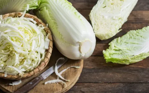 Frischer Chinakohl, bereit für leckere Chinakohl Rezepte. Ein ganzer Kohlkopf, geschnittene Blätter und fein gehackter Kohl in einem Korb liegen auf einem Holztisch.