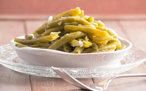 Omas Bohnensalat Rezept: Grüne Bohnen mit Zwiebeln in einer Schüssel auf einem Glasuntersetzer, daneben eine Gabel.