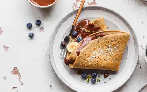 Buchweizen-Crêpes mit Schokoladensoße, Blaubeeren und Banane. Ein köstliches Beispiel für Buchweizen Rezepte, garniert mit Kokosraspeln und Pistazien.