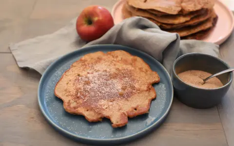 Ein Apfelpfannkuchen, bestreut mit Zucker und Zimt, liegt auf einem blauen Teller. Daneben ein roter Apfel und eine Schüssel mit Zimtzucker. Ein leckeres Apfelpfannkuchen Rezept zum Nachmachen.