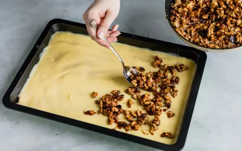 Eine Hand verteilt karamellisierte Walnüsse auf einem hellgelben Teig in einer Backform. Bald gibt es einen "friss dich dumm Kuchen"!
