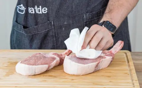 Vorbereitung für ein saftiges Kotelett Rezept: Frische Schweinekoteletts werden auf einem Holzbrett mit Küchenpapier abgetupft.