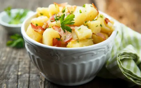 Ein einfacher Kartoffelsalat mit Speck und Petersilie in einer Schüssel, serviert auf einem Holztisch.
