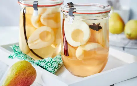 Zwei Gläser mit eingekochten Birnen, verfeinert mit Zimt und Sternanis. Ein frische Birne liegt daneben.