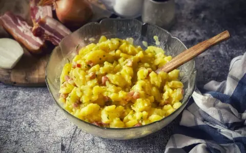 Schüssel mit warmem Kartoffelsalat, garniert mit Speck und Kräutern, daneben Zwiebeln und Speck.