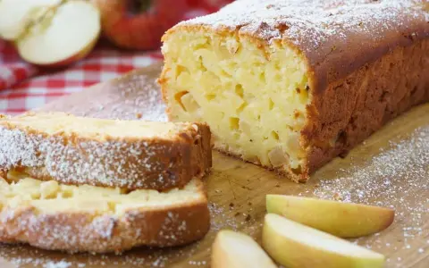 Ein saftiger, schneller Apfelkuchen mit Puderzucker bestäubt, daneben Apfelspalten und ganze Äpfel auf kariertem Tuch.