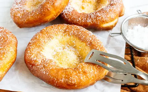 Frisch gebackene, ausgezogene Krapfen, bestäubt mit Puderzucker. Ein Kochlöffel hält einen der goldbraunen Krapfen.
